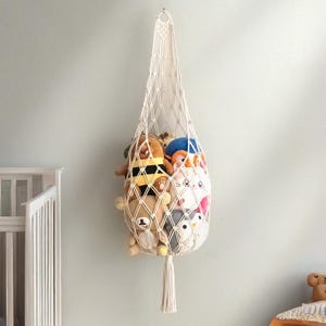 Puede incluir: Una cesta colgante de macramé color crema llena de peluches. La cesta está suspendida de un gancho en una pared azul claro. La cesta tiene un diseño de red en forma de diamante y una borla en la parte inferior. Los peluches incluyen un oso, un pingüino y una abeja.