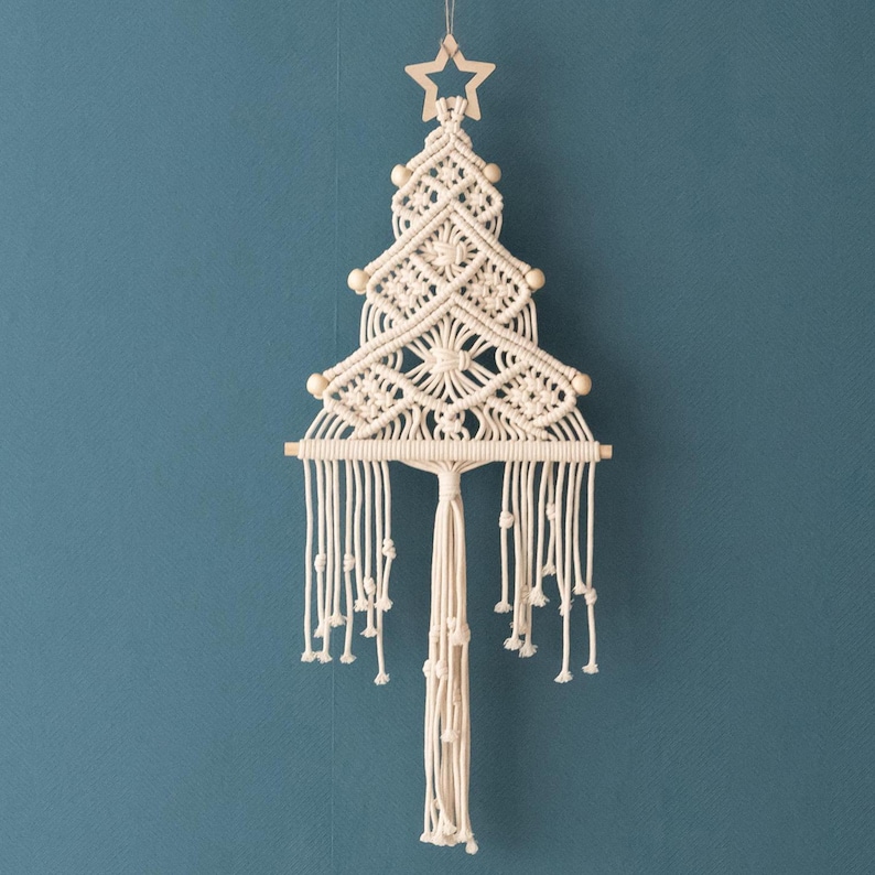May include: A cream-coloured macrame wall hanging shaped like a Christmas tree. The design features intricate knotted patterns, small wooden beads at the branch tips, a wooden star at the top, and a horizontal wooden dowel with long fringe.