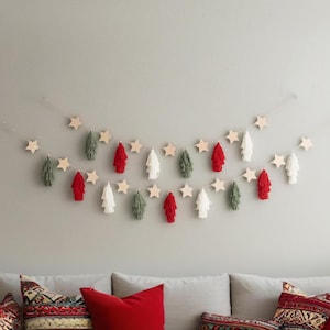Peut inclure: Une guirlande festive avec des étoiles en bois et des pompons colorés rouges, blancs et verts. La guirlande est suspendue sur un mur gris, créant une décoration de vacances. Les pompons sont disposés en motif.