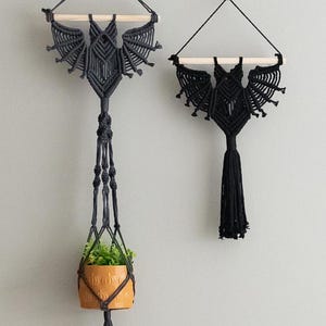Pode incluir: Dois tapetes de parede de macramé preto em forma de morcegos. Um morcego está pendurado em uma vareta de madeira e tem um vaso de planta pendurado nele. O outro morcego está pendurado em uma vareta de madeira e tem uma borla.