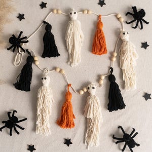 May include: A Halloween garland with handmade ghost figures and tassels in black and orange. The garland is strung with wooden beads and includes black star and spider decorations. Ideal for festive home decor.