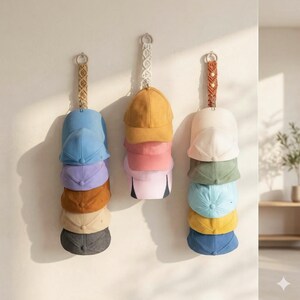 May include: Three wall-mounted hat organisers, each holding a stack of baseball caps in various colours. The caps include shades of blue, purple, brown, and yellow. The organisers have decorative woven straps.