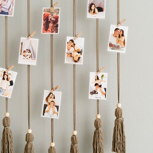 K&ouml;nnte beinhalten: Eine Wanddekoration mit einem Holzstab, an dem braune Schn&uuml;re mit Quasten h&auml;ngen. Fotos sind mit Holz-W&auml;scheklammern an der Schnur befestigt. Die Fotos zeigen verschiedene Personen und Szenen.