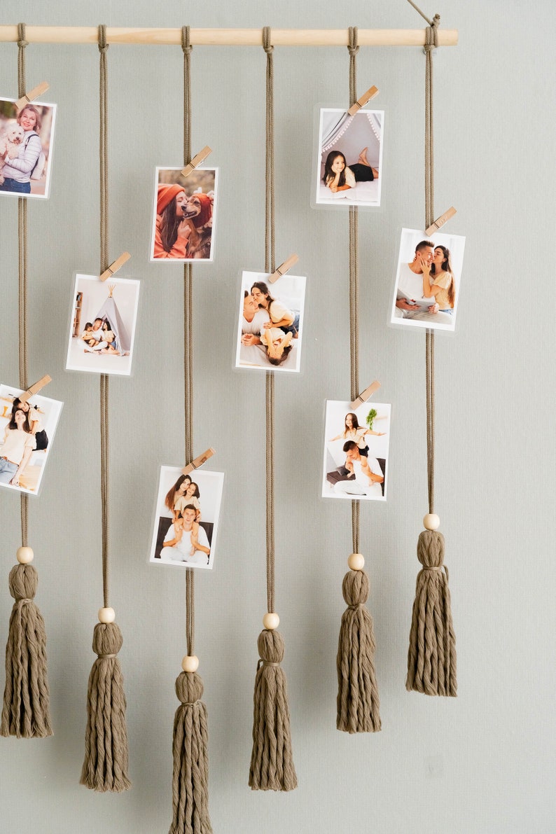 K&ouml;nnte beinhalten: Eine Wanddekoration mit einem Holzstab, an dem braune Schn&uuml;re mit Quasten h&auml;ngen. Fotos sind mit Holz-W&auml;scheklammern an der Schnur befestigt. Die Fotos zeigen verschiedene Personen und Szenen.