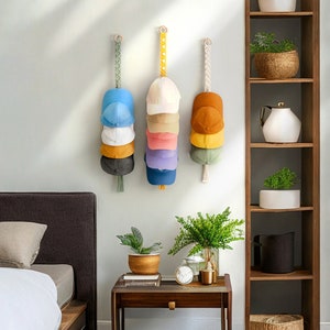 May include: Three wall-mounted hat organizers, each holding a stack of baseball caps in various colors. The organizers are suspended by decorative straps with wooden rings. The caps include shades of blue, white, yellow, pink, and green.