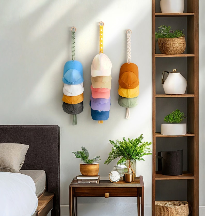 May include: Three wall-mounted hat organizers, each holding a stack of baseball caps in various colors. The organizers are suspended by decorative straps with wooden rings. The caps include shades of blue, white, yellow, pink, and green.