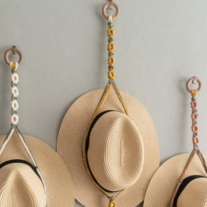 May include: Three straw hats suspended from a wall by macrame hangers. Each hanger has a wooden ring and a daisy chain in colours like yellow, white, and pink. The hats have black bands. The hangers are decorative.