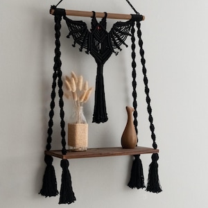 May include: A black macrame wall shelf with a wooden shelf and a decorative macrame design. The shelf is suspended by black rope and features a wooden rod at the top. The shelf is decorated with a glass vase, dried flowers, and a brown vase.