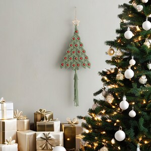 K&ouml;nnte beinhalten: Ein gr&uuml;ner Makramee-Weihnachtsbaum-Wandbehang mit roten Perlen und einem Holzstern an der Spitze. Der Baum hat eine lange gr&uuml;ne Quaste, die von unten h&auml;ngt.
