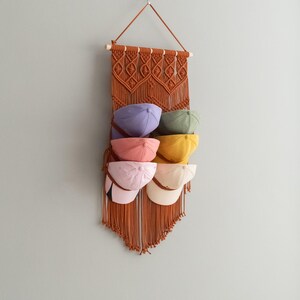 May include: A macrame wall hanging with a brown and beige color scheme. The hanging has six pockets for storing hats. The hats are in various colors, including pink, purple, yellow, and green.