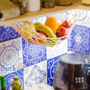 May include: A white string hammock hanging from a wooden shelf, filled with fruit, including oranges, apples, bananas, and a green pear. The hammock is hanging over a blue and white patterned tile backsplash.