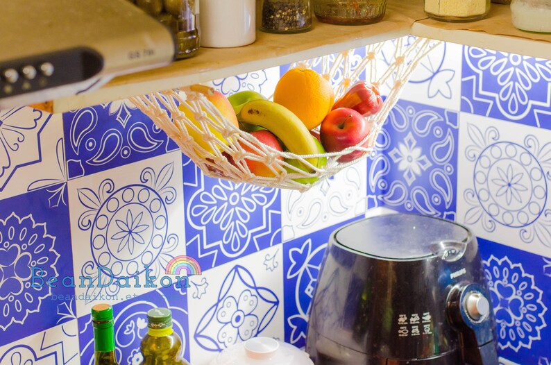May include: A white string hammock hanging from a wooden shelf, filled with fruit, including oranges, apples, bananas, and a green pear. The hammock is hanging over a blue and white patterned tile backsplash.