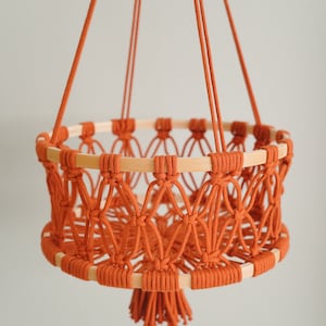 May include: A hanging macrame basket with a wooden ring and a tassel. The basket is made of orange cord and is hanging from a white wall.