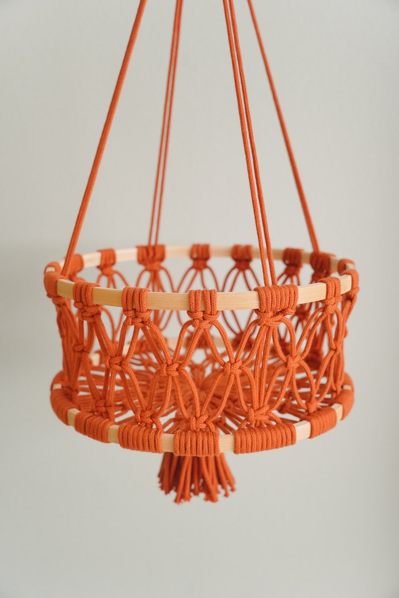 May include: A hanging macrame basket with a wooden ring and a tassel. The basket is made of orange cord and is hanging from a white wall.