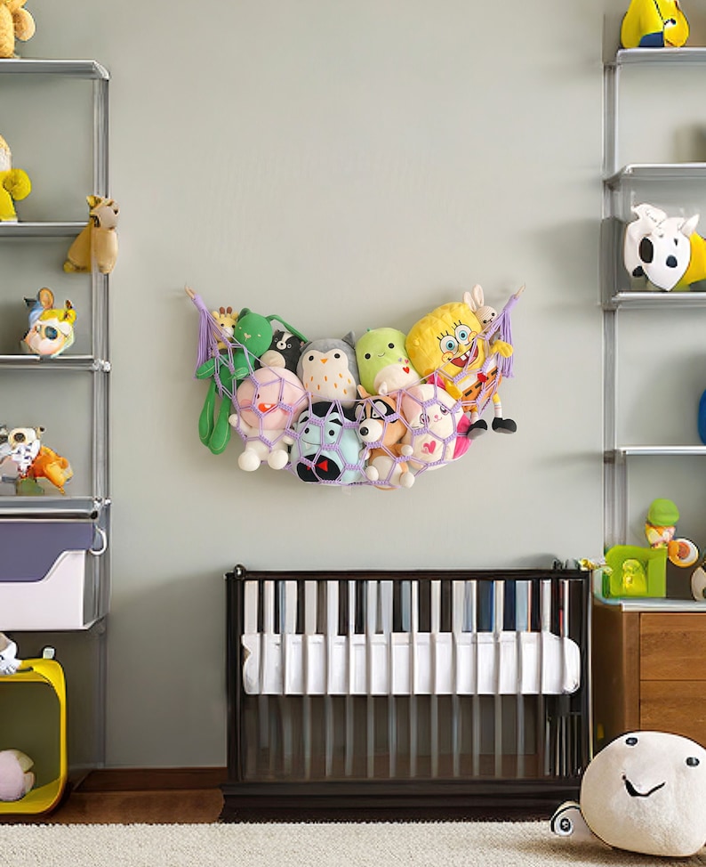 Op de afbeelding: Een paarse en witte net-hangmat gevuld met knuffels, hangend aan een lichtgroene muur. De hangmat is gevuld met een verscheidenheid aan knuffels, waaronder een groene kikker, een zwart-witte wasbeer, een roze varken, een witte uil, een groene kikker, een gele spons, een wit konijn en een roze kat. De hangmat hangt boven een witte wieg met een donkerbruin frame.
