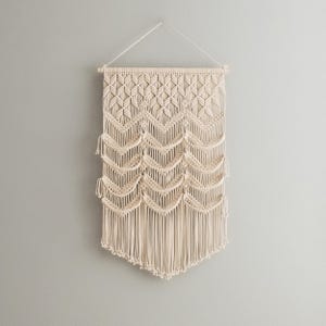May include: A beige macrame wall hanging with a geometric pattern and fringe. The hanging is made of natural fibers and has a bohemian style.