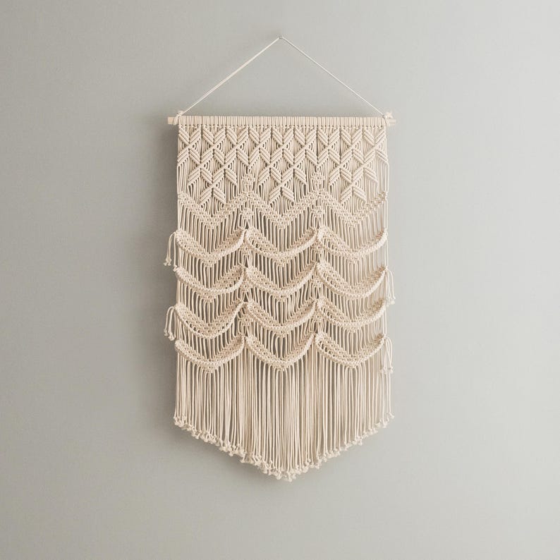 May include: A beige macrame wall hanging with a geometric pattern and fringe. The hanging is made of natural fibers and has a bohemian style.