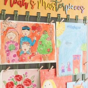 Puede incluir: Un expositor de pared de madera con las palabras "Noah's Masterpieces" en letras de colores. El expositor tiene cuerda verde y pinzas de madera que sujetan dibujos coloridos de personas, flores y una casa.