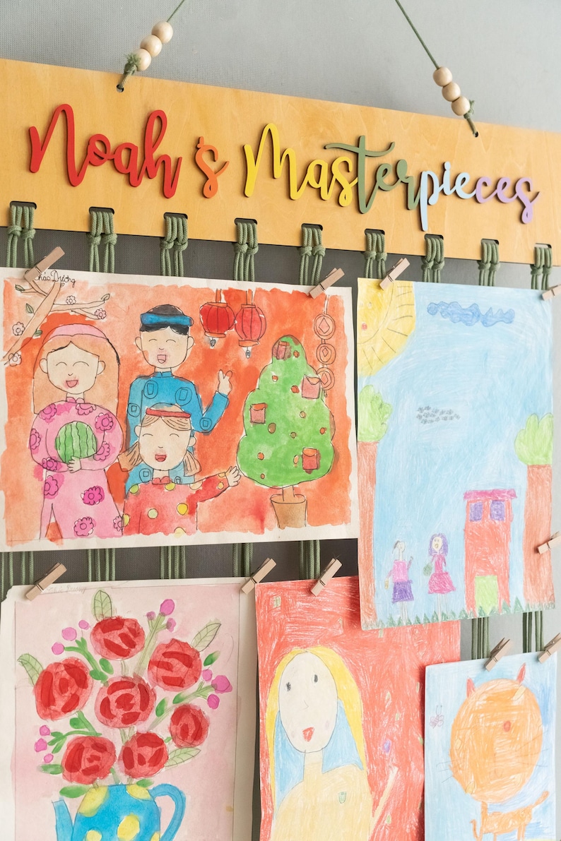 Puede incluir: Un expositor de pared de madera con las palabras "Noah's Masterpieces" en letras de colores. El expositor tiene cuerda verde y pinzas de madera que sujetan dibujos coloridos de personas, flores y una casa.