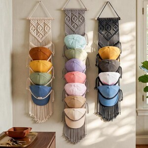 May include: Three macrame hat organisers in cream, grey, and black, each holding baseball caps in various colours. The organisers hang from wooden dowels, with a woven design at the top and fringe at the bottom. The hats are in shades of brown, yellow, green, and blue.