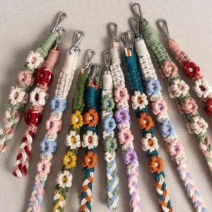 May include: A collection of braided macrame wrist straps in various colors, each adorned with small, colorful flower accents. The straps feature silver-tone metal clasps for easy attachment. The color palette includes pink, red, blue, green, and orange.