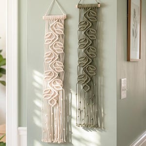 Puede incluir: Dos tapices de macramé verticales colgados de varillas de madera. Una pieza es de color crema y la otra verde oliva. Ambas presentan intrincados patrones de nudos que forman hojas, con largos flecos colgantes en los extremos inferiores.