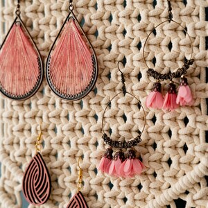 May include: A close-up of a macrame wall hanging with various earrings hanging from it. The earrings are made of different materials, including wood, metal, and fabric. The earrings are all in shades of pink and brown.
