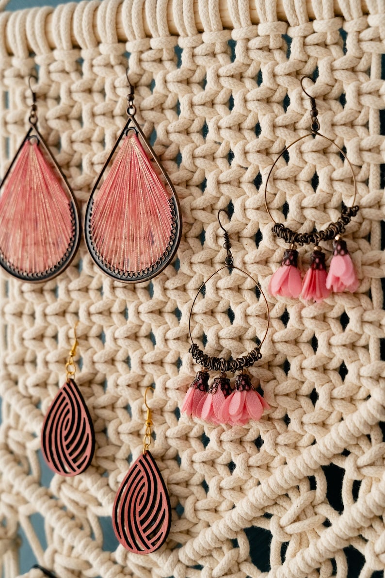 May include: A close-up of a macrame wall hanging with various earrings hanging from it. The earrings are made of different materials, including wood, metal, and fabric. The earrings are all in shades of pink and brown.