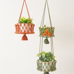 May include: Two macrame plant hangers with wooden rings. The top hanger is orange and holds a potted plant. The bottom hanger is green and has two tiers. The top tier holds oranges and the bottom tier holds a potted plant.