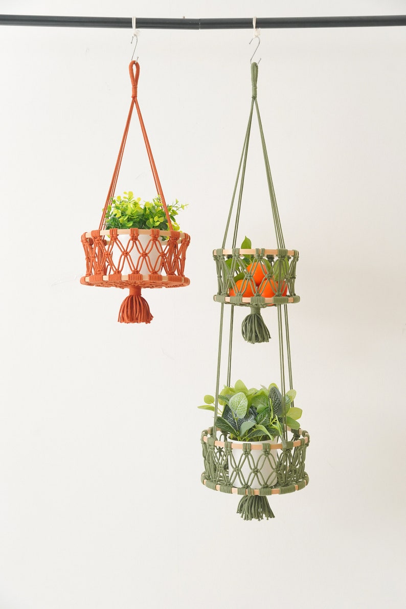 May include: Two macrame plant hangers with wooden rings. The top hanger is orange and holds a potted plant. The bottom hanger is green and has two tiers. The top tier holds oranges and the bottom tier holds a potted plant.