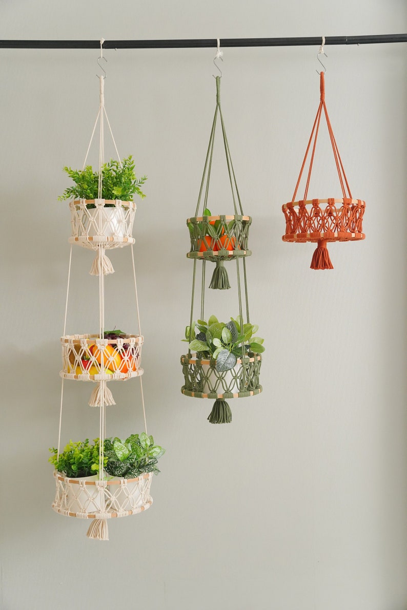 May include: Three macrame plant hangers with wooden rings and tassels. The top hanger is white and has three tiers, the middle hanger is green and has two tiers, and the bottom hanger is orange and has one tier.