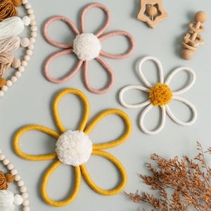 May include: Three handmade flower decorations in pink, white, and yellow, each with a fluffy white pom-pom center. The flowers are made of looped yarn. Additional wooden toys and decorative beads are also visible.