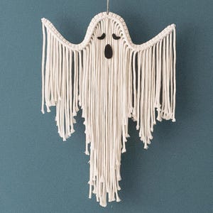 Puede incluir: Un fantasma de macramé blanco para colgar en la pared con una cara negra. El fantasma está hecho de hilo y tiene flecos alrededor de los bordes.