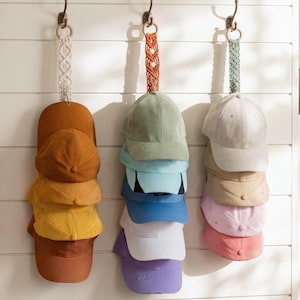 Può includere: Tre set di cappellini da baseball appesi a una parete bianca. Ogni set è sospeso a un gancio decorativo. I cappellini sono in vari colori, tra cui marrone, giallo, verde, blu, bianco e rosa. I cinturini sono bianchi, arancioni e verdi.
