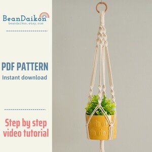 Puede incluir: Un macramé blanco para colgar plantas con una maceta amarilla y una planta verde. El macramé está hecho con un patrón de nudos y tiene un anillo de madera en la parte superior. El texto "PDF PATTERN Instant download" y "Step by step video tutorial" es visible en la imagen.