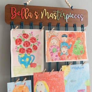 Puede incluir: Un letrero de madera con el texto "Bella's Masterpieces" cuelga de una pared.  El letrero tiene una cuerda azul con cuentas de madera.  La cuerda se utiliza para colgar obras de arte en la pared.  Hay cinco obras de arte colgadas de la cuerda.  Las obras de arte son coloridas e incluyen dibujos de flores, personas y un gato.