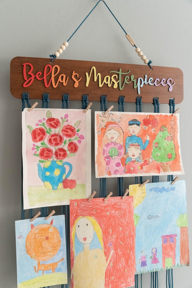 Puede incluir: Un letrero de madera con el texto "Bella's Masterpieces" cuelga de una pared.  El letrero tiene una cuerda azul con cuentas de madera.  La cuerda se utiliza para colgar obras de arte en la pared.  Hay cinco obras de arte colgadas de la cuerda.  Las obras de arte son coloridas e incluyen dibujos de flores, personas y un gato.