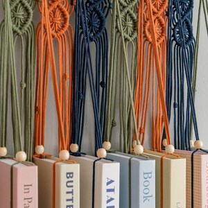 May include: A wall hanging made of macrame with different coloured cords in green, orange, and blue. The macrame is hanging over a row of books with titles like "Butter", "A Table in the...