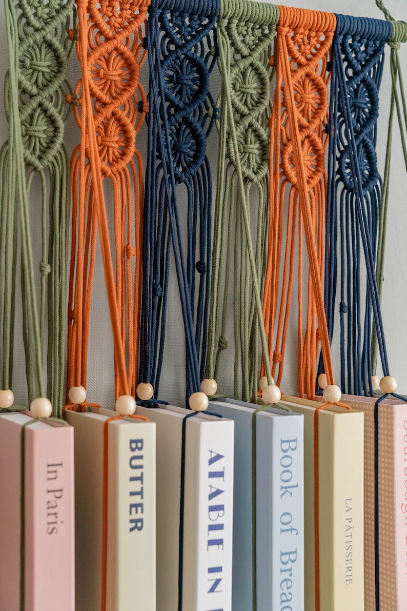 May include: A wall hanging made of macrame with different coloured cords in green, orange, and blue. The macrame is hanging over a row of books with titles like "Butter", "A Table in the...