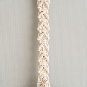 May include: Cream-colored macrame plant hanger with a wooden ring at the top and a metal clip at the bottom. The macrame design features a series of diamond-shaped knots. The bottom clip holds a brown, rounded object.