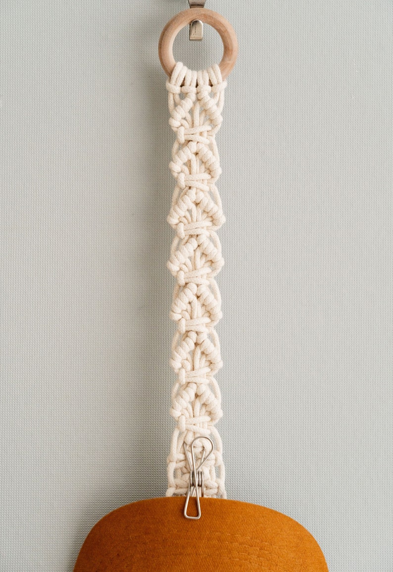 May include: Cream-colored macrame plant hanger with a wooden ring at the top and a metal clip at the bottom. The macrame design features a series of diamond-shaped knots. The bottom clip holds a brown, rounded object.