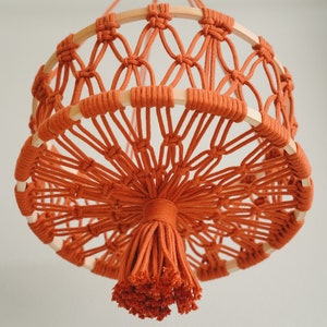 May include: A macrame hanging lamp with a round, woven base and a tassel made of orange cord.