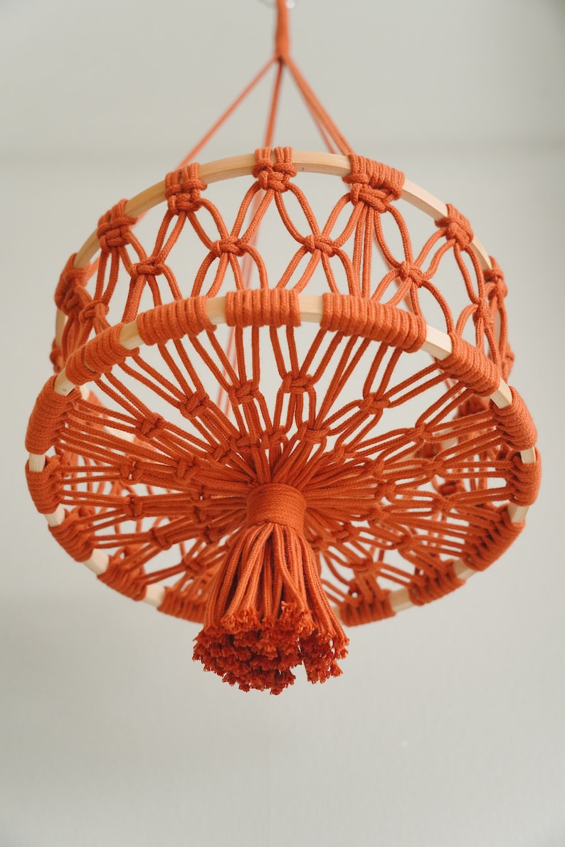 May include: A macrame hanging lamp with a round, woven base and a tassel made of orange cord.
