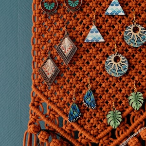 May include: A macrame wall hanging with a wooden dowel at the top and tassels at the bottom. The wall hanging is made of brown cord and has various earrings hanging from it. The earrings are made of different materials, including wood, metal, and glass. Some of the earrings are shaped like butterflies, leaves, and geometric shapes. The earrings are decorated with various colors and patterns.