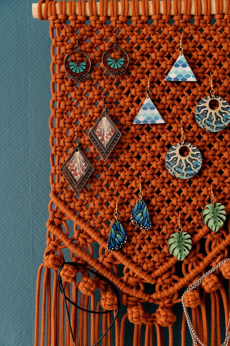 May include: A macrame wall hanging with a wooden dowel at the top and tassels at the bottom. The wall hanging is made of brown cord and has various earrings hanging from it. The earrings are made of different materials, including wood, metal, and glass. Some of the earrings are shaped like butterflies, leaves, and geometric shapes. The earrings are decorated with various colors and patterns.