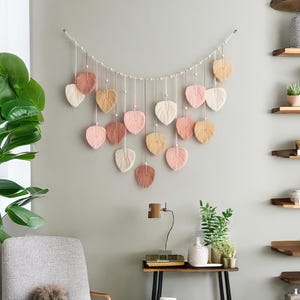 K&ouml;nnte beinhalten: Ein Makramee-Wandbehang mit einer Schnur aus rosa, pfirsichfarbenen und cremefarbenen Bl&auml;ttern. Die Bl&auml;tter sind mit wei&szlig;en Perlen aufgereiht und h&auml;ngen an einer wei&szlig;en Schnur.