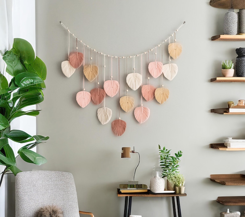 K&ouml;nnte beinhalten: Ein Makramee-Wandbehang mit einer Schnur aus rosa, pfirsichfarbenen und cremefarbenen Bl&auml;ttern. Die Bl&auml;tter sind mit wei&szlig;en Perlen aufgereiht und h&auml;ngen an einer wei&szlig;en Schnur.