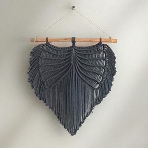 Könnte beinhalten: Dunkelgrauer Makramee-Wandbehang in Blattform. Das Makramee ist an einem natürlichen Holzstab aufgehängt. Das Blattdesign weist ein zentrales Adernmuster und gefranste Kanten auf, wodurch ein strukturiertes, dekoratives Stück entsteht.