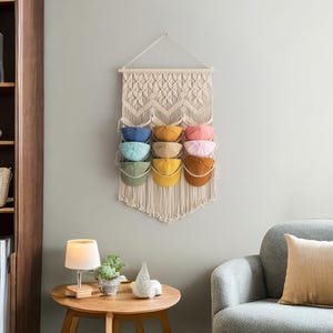 May include: A macrame wall hanging with a geometric pattern, featuring eight slots for storing hats. The slots are filled with colorful hats in shades of blue, green, yellow, orange, and pink.
