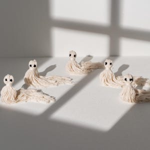 May include: Five white cotton tassels with black eyes, arranged in a row on a white surface. The tassels are handmade and have a soft, fluffy texture.